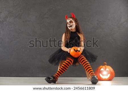 Happy child girl with festive makeup in carnival black costume of devil with horns sitting in the studio on gray background with copy space holding orange Halloween pumpkin and laughing.