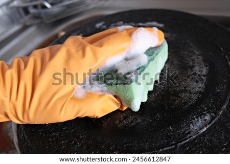 A man's hand in a protective glove cleans a pan with a burnt bottom with a foam kitchen sponge. Royalty-Free Stock Photo #2456612847