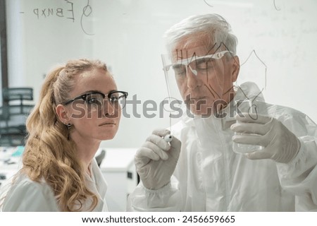 Two chemist colleagues write formulas on glass. Caucasian elderly man and young woman brainstorming.