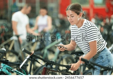 Interested young girl using smartphone to photograph bicycle in shop, to share with friends for opinions or to compare with other options online