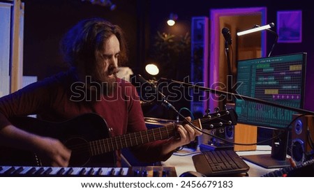 Creative songwriter singing a song on microphone and playing his guitar, acoustical engineering technology with daw software on pc. Artist creating music with instrument and mixing console. Camera B.
