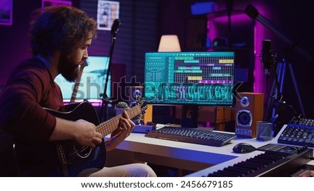 Musical performer recording guitar sounds for a new track in home studio, soundboard controls and daw software on computer. Young musician playing acoustic instrument, music industry. Camera B.