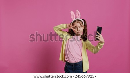 Confident happy young girl taking photos on smartphone webcam, feeling cute with her pigtails and bunny ears. Joyful smiling toddler takes pictures and fooling around in studio. Camera B.