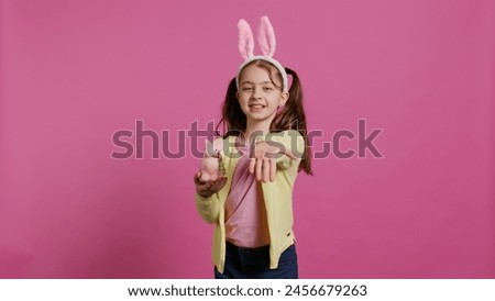 Lovely little girl with bunny ears sending air kisses in studio, presenting a stuffed pink rabbit toy for easter holiday. Smiling energetic preteen feeling positive and carefree. Camera B.