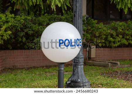 An illuminated round white globe made of glass with blue lettering. The police sign is on a black aluminum cast base. The background is grass, trees, and a wooden fence. A sign of a police station.