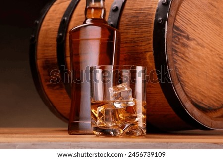 Whiskey with ice cubes in glass, bottle and barrel on wooden table against dark background, closeup