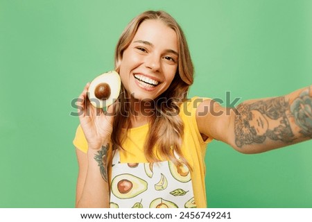 Close up young housewife housekeeper chef cook baker woman wear apron yellow t-shirt hold avocado do selfie shot on mobile cell phone isolated on plain pastel green background. Cooking food concept