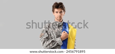 Young male Ukrainian soldier with flag on grey background  