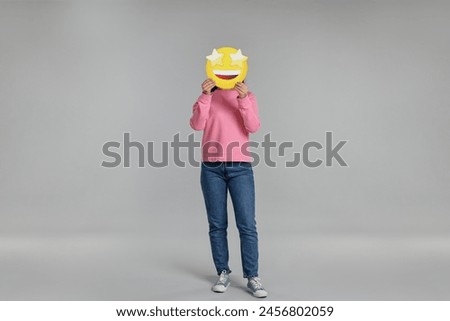 Woman holding emoticon with stars instead of eyes on grey background