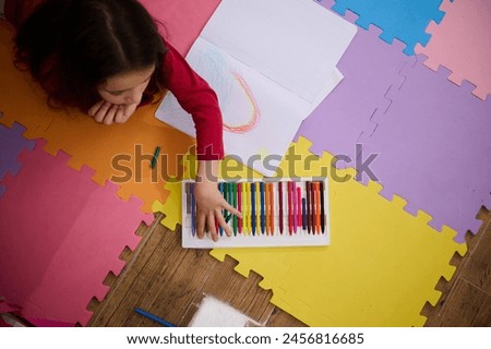 View from above of a little child girl taking out colorful pencil from pencil case, drawing beautiful cloud with rainbow, lying on a multi colored puzzle carpet in cozy home interior. Kids education