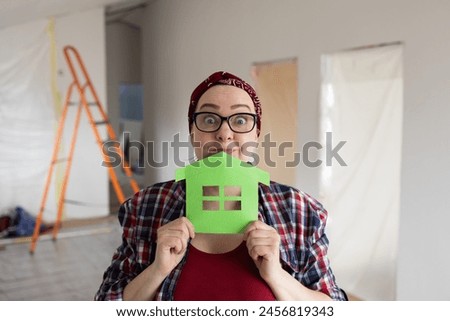 A joyful woman with surprised eyebrows holds a paper picture near her face, a symbol of an eco-friendly home. High quality photo