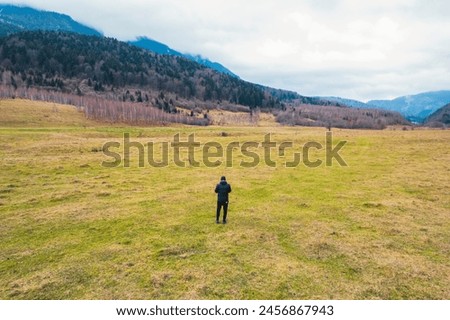 A person stands gracefully in a vast field, surrounded by majestic mountains in the distant background