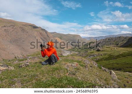 Photographer in vivid red wind jacket with hood and large camera in hands in high mountains. Guy on grassy hill takes pictures in alpine valley in sunny day. Clouds in blue sky in changeable weather.