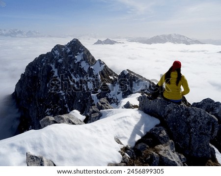 Mujer de espaldas en lo alto de una montaña nevada, sobre un mar de nubes con un espolón rocoso y montañas en el horizonte con cielo azul. Royalty-Free Stock Photo #2456899305