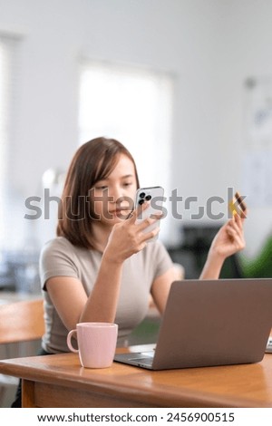 A woman is sitting at a desk with a laptop and a cell phone. She is looking at her cell phone and holding a credit card