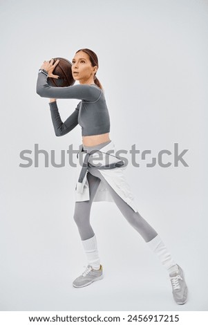 Active young woman in sportswear confidently holds a basketball in her right hand against a grey background.