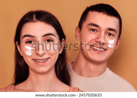 Close-up of teenagers with cream on their cheeks. A cute smiling girl with braces and a funny guy take care of the skin of the face. Hygiene and skin care in adolescence. Isolated beige background.