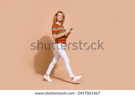 Full size photo of positive cheerful girl wear ornament t-shirt walk with smartphone in empty space isolated on beige color background