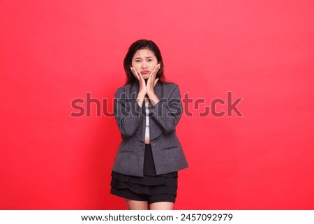 gesture of an Asian office woman pouting with both hands holding her cheeks wearing a gray jacket and mini skirt on a red background. for fashion, lifestyle and business concepts