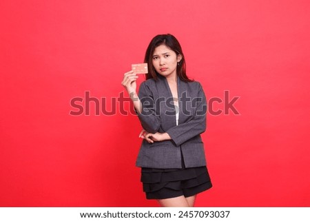 Expression of astonished chinese businesswoman with arms crossed and holding a debit credit card wearing a jacket and skirt on a red background. for financial, business and advertising concepts