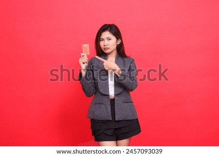 sad indonesia businesswoman expression into camera hand holding debit credit card while pointing at it wearing jacket and skirt on red background. for financial, business and advertising concepts