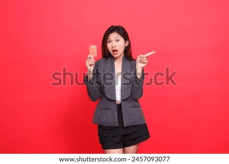 Asian office girl expression shocked, holding a debit credit card while pointing to the left, wearing a jacket and skirt on a red background. for financial, business and advertising concepts