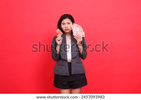 gesture of sad indonesia office woman holding credit card and money in head area wearing jacket and skirt on red background. for transaction, business and advertising concepts