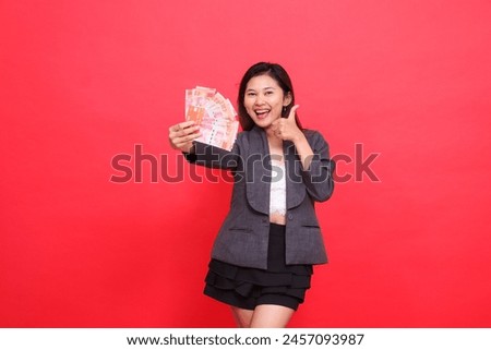 gesture of a happy Asian career woman holding a credit card and money in front of a thumbs up sign wearing a jacket and skirt on a red background. for transaction, business and advertising concepts