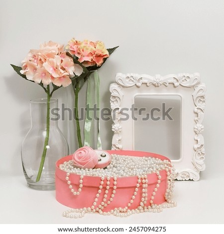 A still life of pink flowers in a glass vase, a picture frame, and a jewelry box with pearls. Hydrangeas in full bloom, a white frame, and a pink round jewelry box filled with pearls.