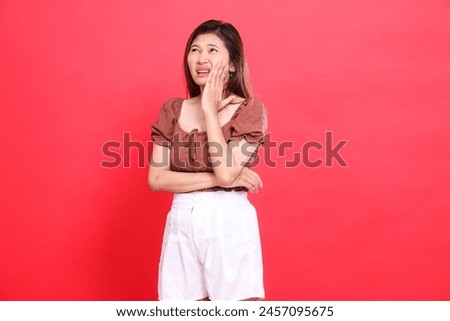 indonesia woman's gesture, candid, crooked to the right, holding her cheek, toothache, pain, wearing a brown blouse and shorts with a red background. for health, care and advertising concept