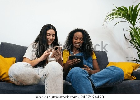 This stock photo features two young women at home, effortlessly using mobile devices and showcasing a modern, connected lifestyle