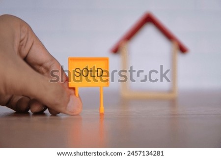 A close-up photo of a hand holding a bright orange "SOLD" sign in front of a miniature wooden house. Real estate and property concept.