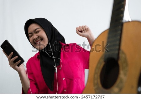 Portrait of an acoustic guitar next to a young Asian Muslim woman in hijab listening to music on her smartphone while singing, focus selected, isolated against a white background.