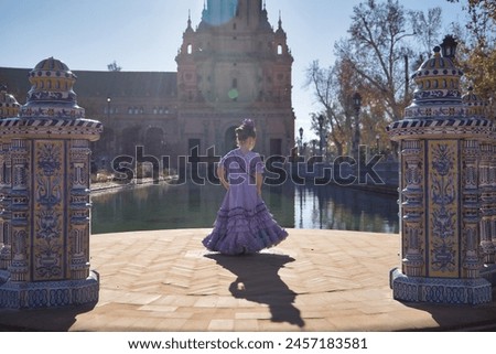 A girl dancing flamenco, in typical flamenco dress, with her back turned, receiving the sun's rays, next to a lake in a beautiful square in Seville. Concept dance, flamenco, typical Spanish, Spain. Royalty-Free Stock Photo #2457183581