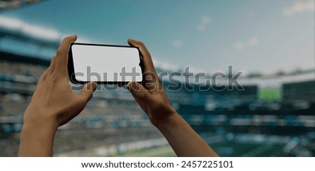Image of a male hand holding a smartphone with a blank screen at an American football stadium. Ideal for showcasing apps related to sports betting, live streaming, and event analytics