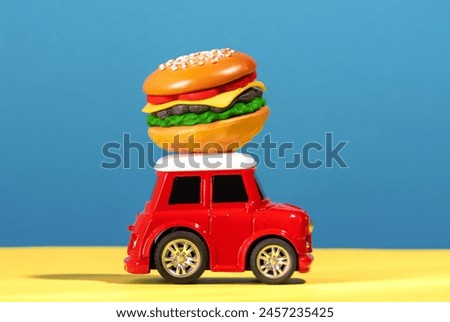 Small red car carries burger on roof on blue and yellow background.