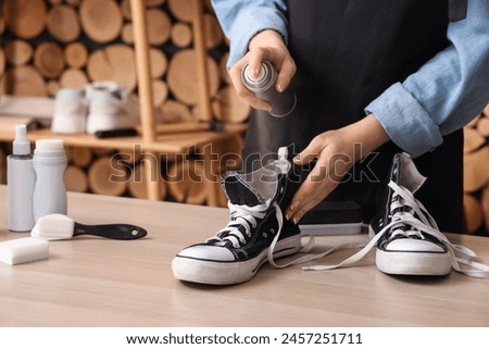 Female shoemaker applying water repellent spray over pair of gumshoes at wooden table, closeup Royalty-Free Stock Photo #2457251711