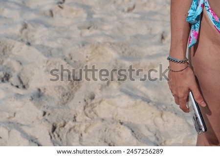 A female arm and hand holding a cell phone, wearing a blue white and pink bikini, wearing a bracelet, standing on the beach, on the white sand on a sunny day