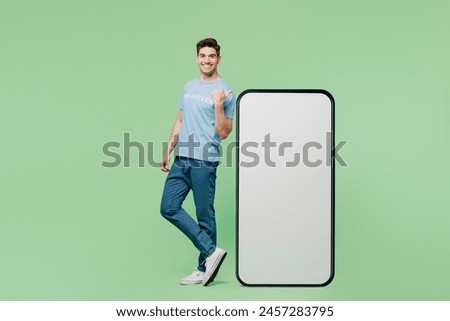 Full body young man wear blue t-shirt title volunteer point finger on big huge blank screen area mobile cell phone isolated on plain green background. Voluntary free work help charity grace concept
