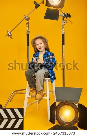 Creative film professions. A stylish little girl sits on the set surrounded by spotlights with an old camera in her hands and is smiling. Full-length portrait on a yellow background.