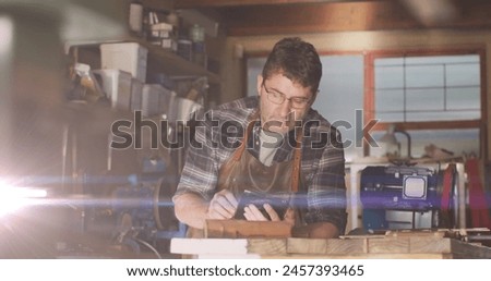 Image of glowing light over caucasian man using tablet in workshop. labor day, work, workers, tradition and celebration concept digitally generated image.