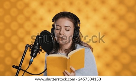 Narrator wearing headset reading aloud from book into mic against yellow background. Upbeat professional voice actor recording audiobook, creating engaging media content for listeners, camera A