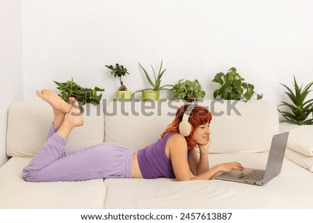 A woman is laying on a couch with a laptop in front of her. She is wearing headphones and she is listening to music while working on her laptop. The room is decorated with potted plants