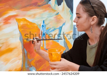 Attractive female painter passionately draws picture with paintbrush for outdoor street exhibition, close up side view of female artist infusing life into outdoor art space