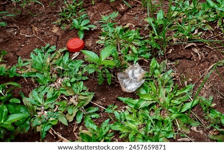Plastic material trash garbage junk. Red colored bottle cap lid and small plastic bag on forest or garden soil ground area with green grass and leaves. Royalty-Free Stock Photo #2457659871
