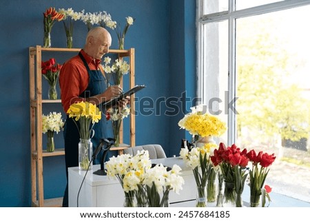 An elderly man flower seller writes with a blue pen on paper with concentration. In front of him is a window overlooking the street