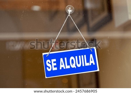 Blue sign hanging at the glass door of a shop saying in Spanish "Se alquila" meaning in English "To rent".
