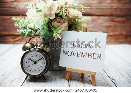 Happy November text message written on paper card with wooden easel and alarm clock with flower in metal vase decoration