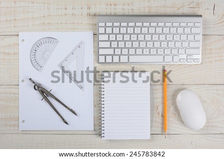 Overhead shot of a computer keyboard and mouse next to a sheet of graph paper, a compass and protractor, on a whitewashed wood table, in a home office.