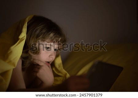 Kid boy lying on bed and surfing Internet on tablet in dark room. Child using tablet pc at night. Little kid in bed under a blanket looking at the smartphone or tablet at night. Gadget addicted child.